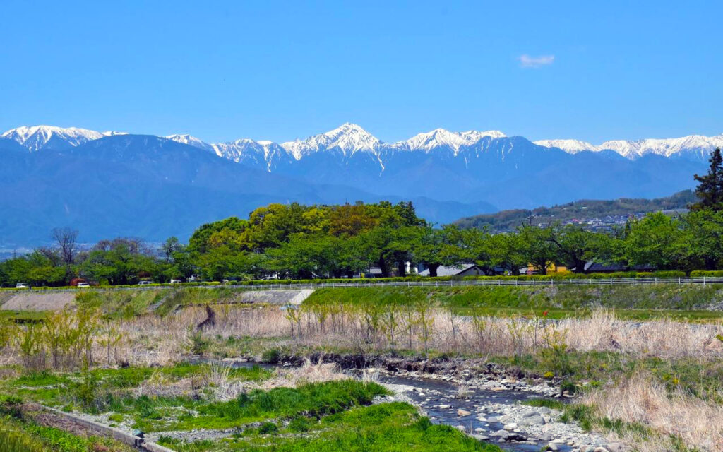 青空の下に広がる雪を頂いた山脈と、手前の小川や新緑の木々。